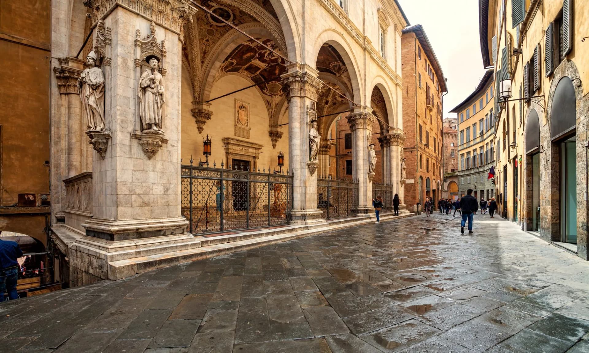 Wet cobblestone street in Siena, Italy, beside a historic building with statues and arches.