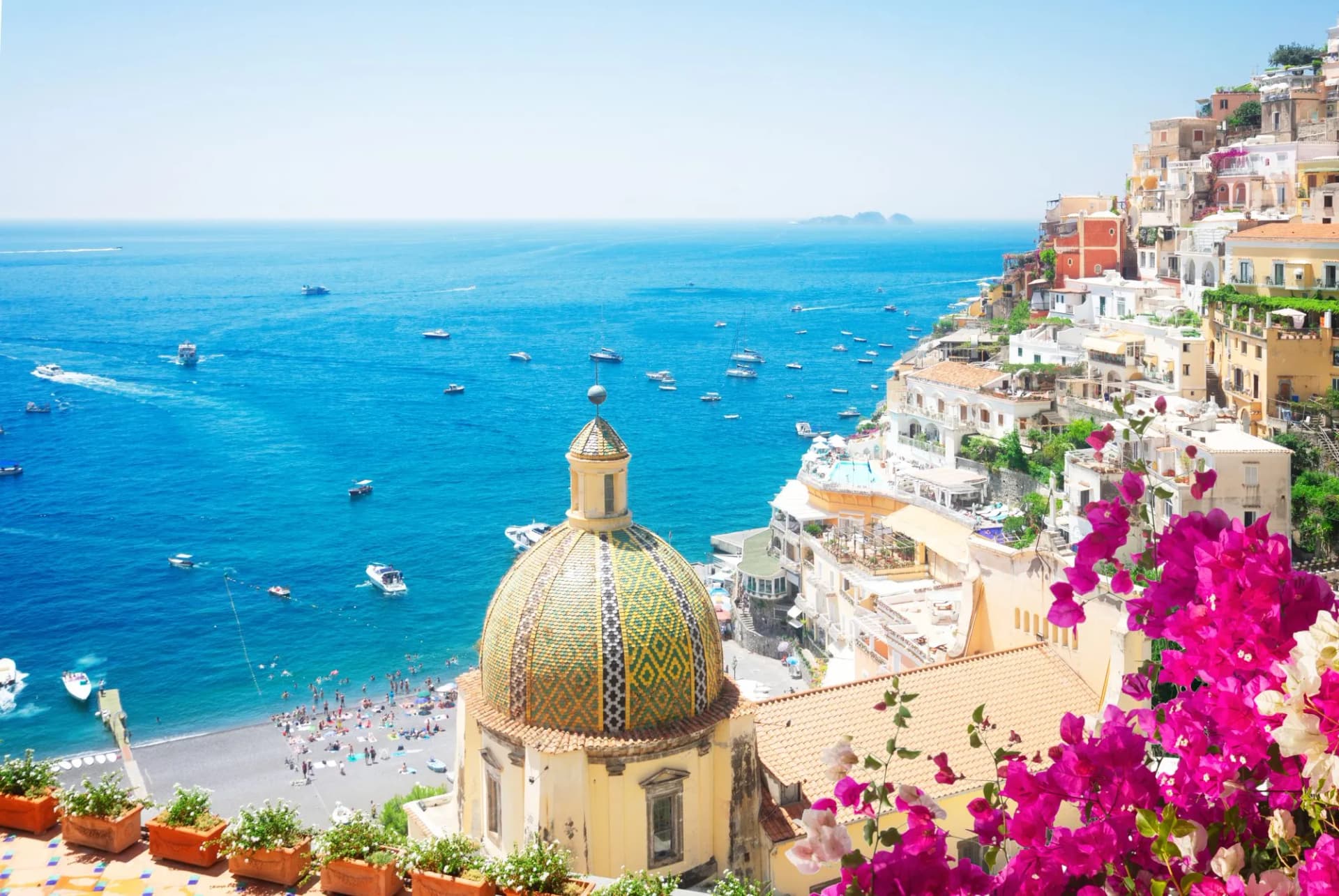 Positano with tiled dome church, colorful cliffside buildings, and boats on blue sea.