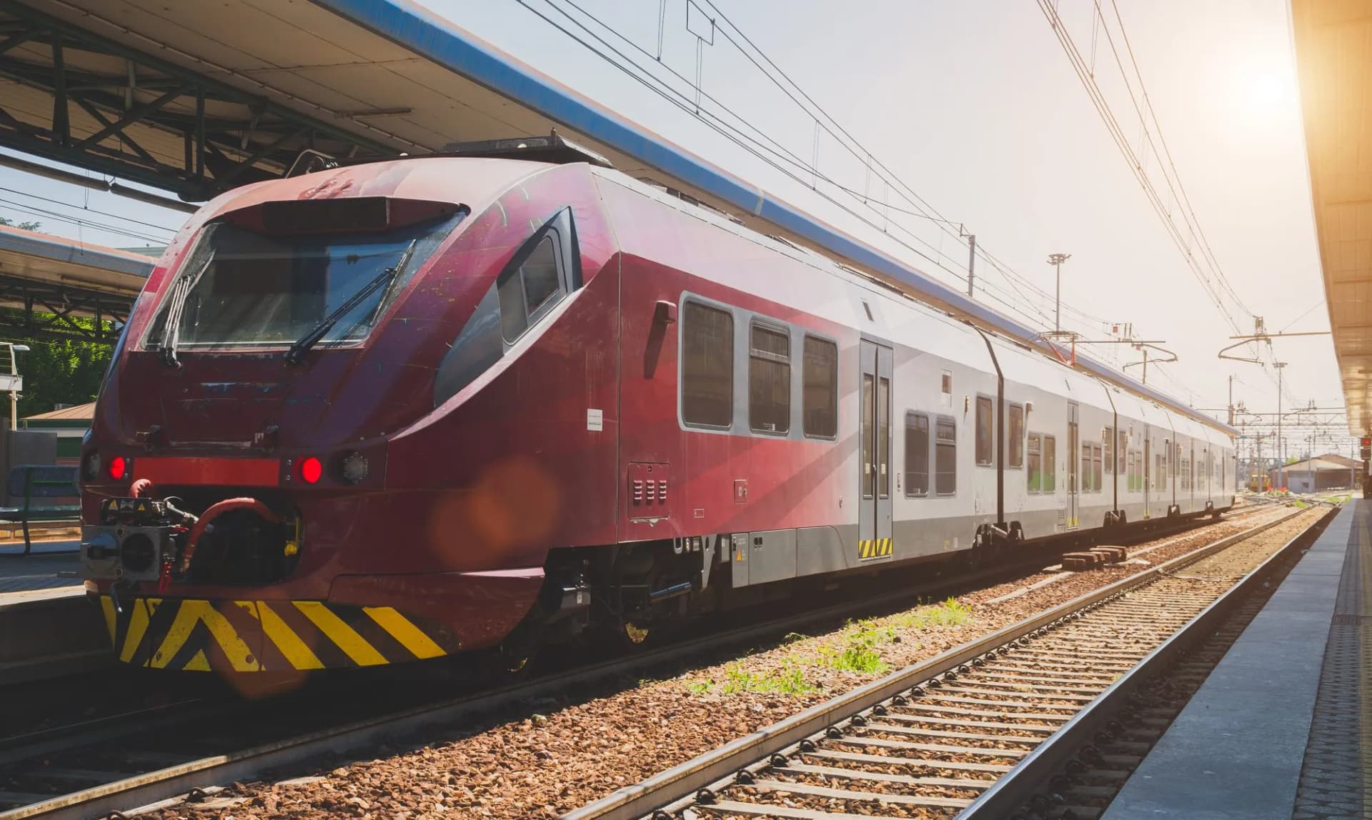 Red and white regional train stopped at a sunny railroad station platform in Northern Italy.