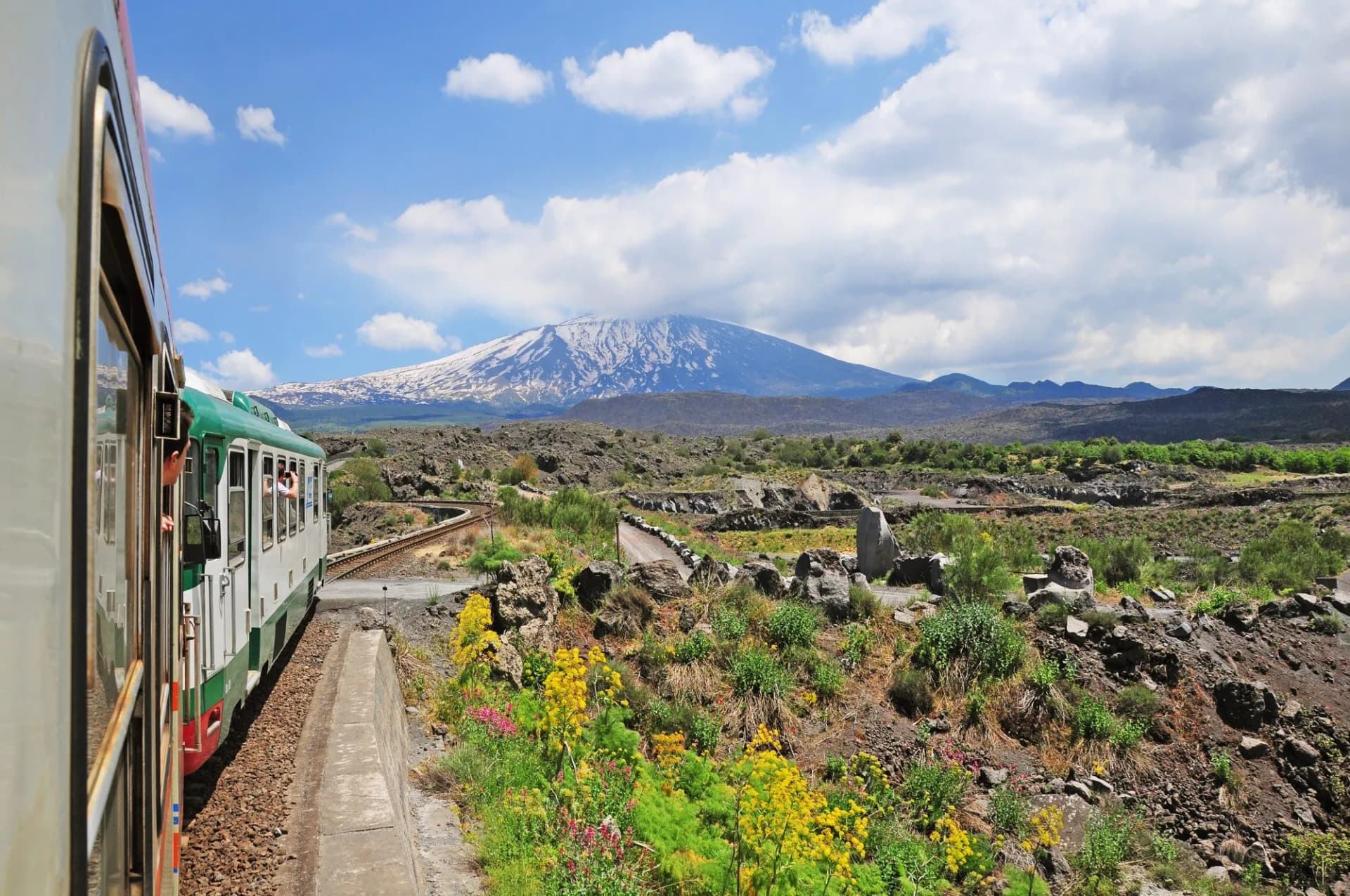 Train traveling past Mount Etna with snow-capped peak through volcanic landscape in Sicily