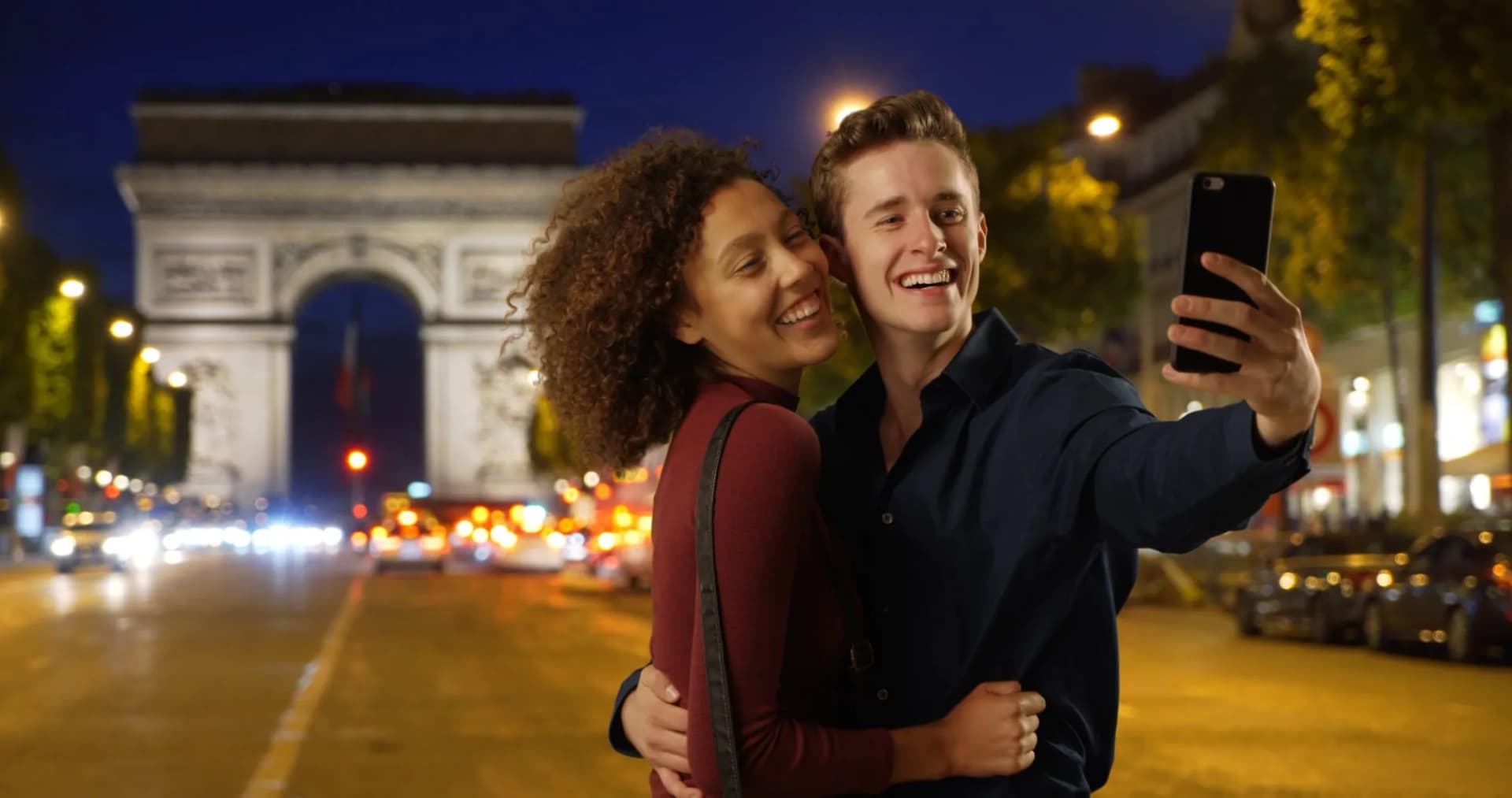 Couple taking a selfie at night on a Paris street with the Arc de Triomphe illuminated.