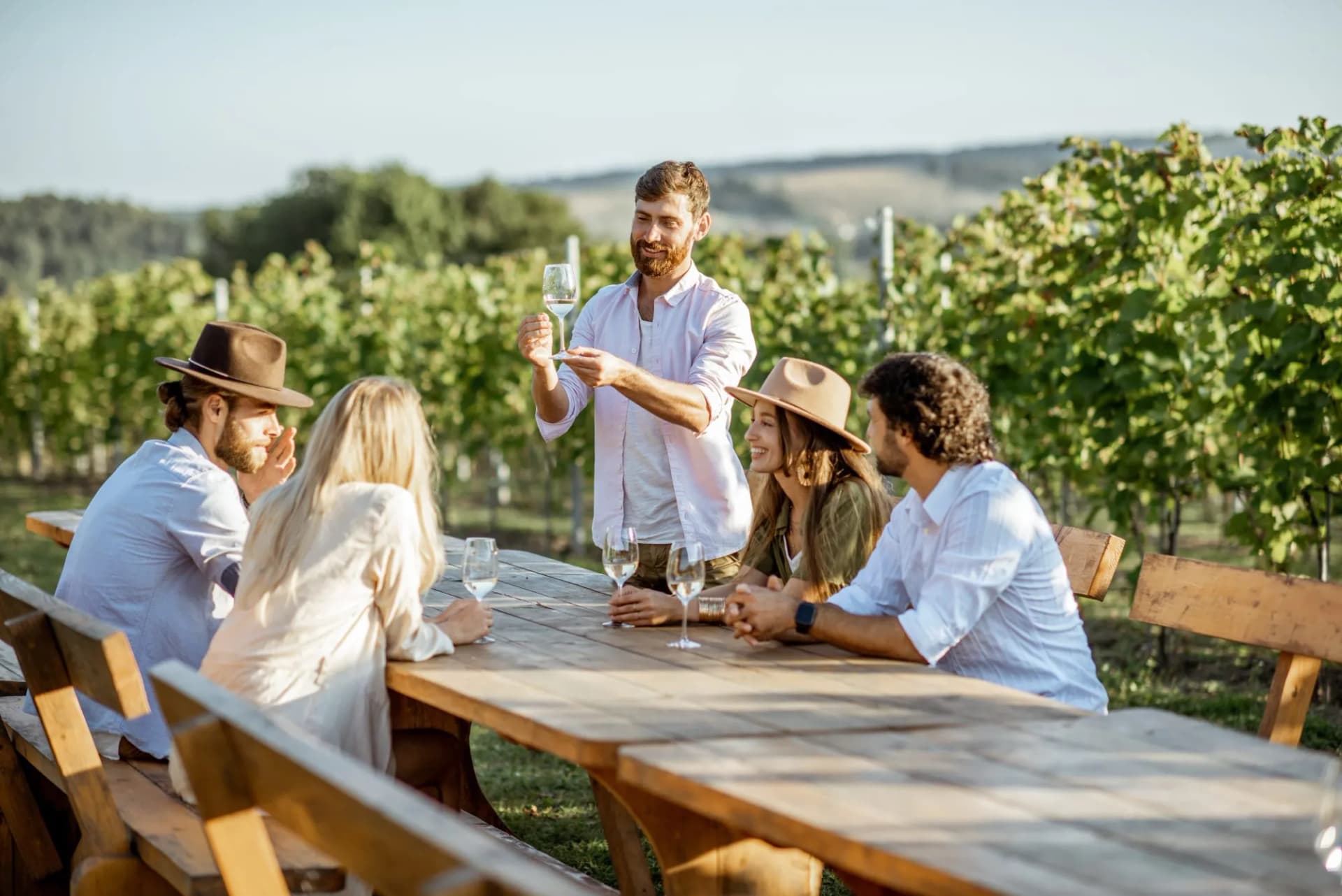 Group tasting wine while sitting at a wooden table outdoors in a sunny vineyard setting.