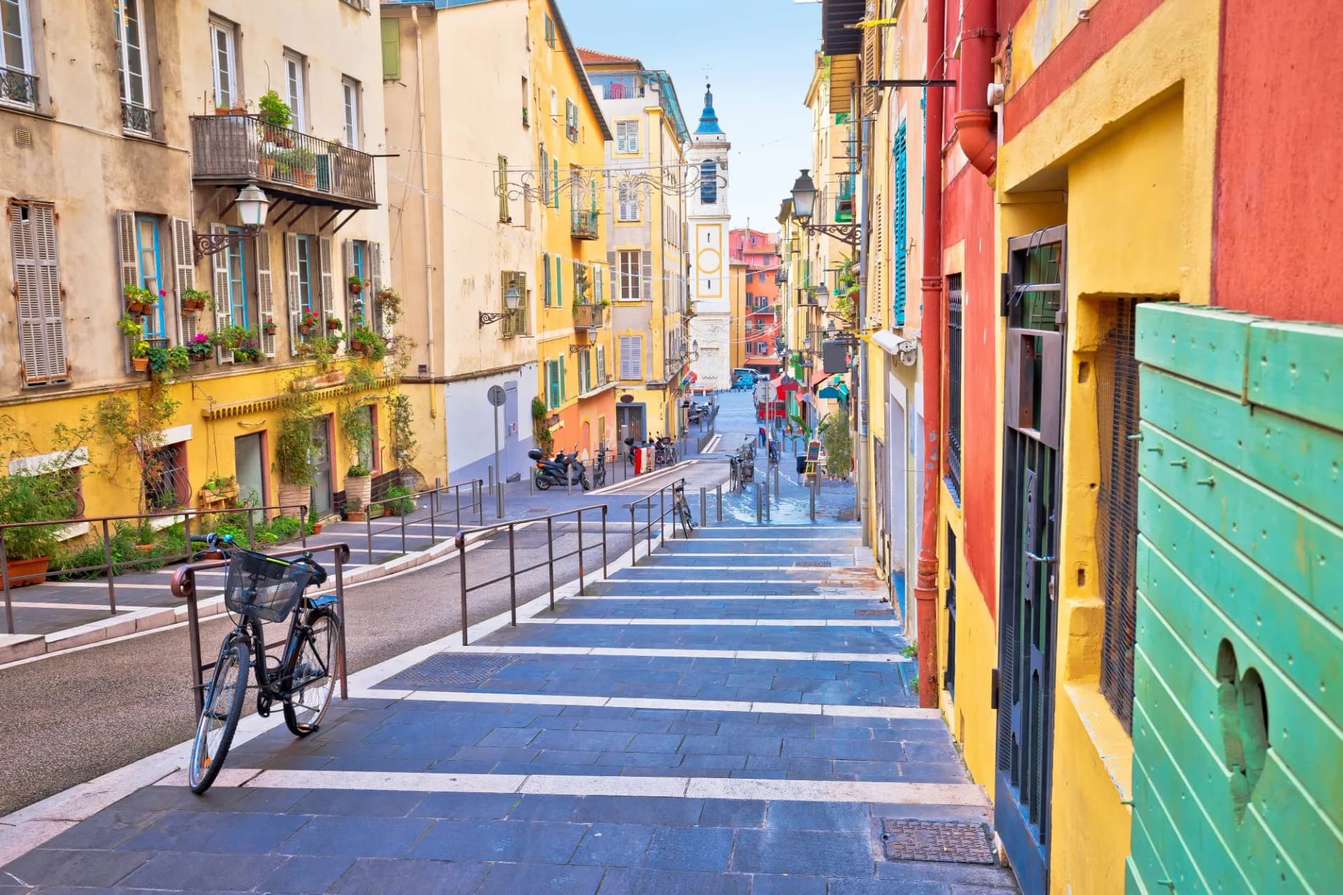 Colorful street in Nice with stairs, a bicycle, and a distant church tower view.