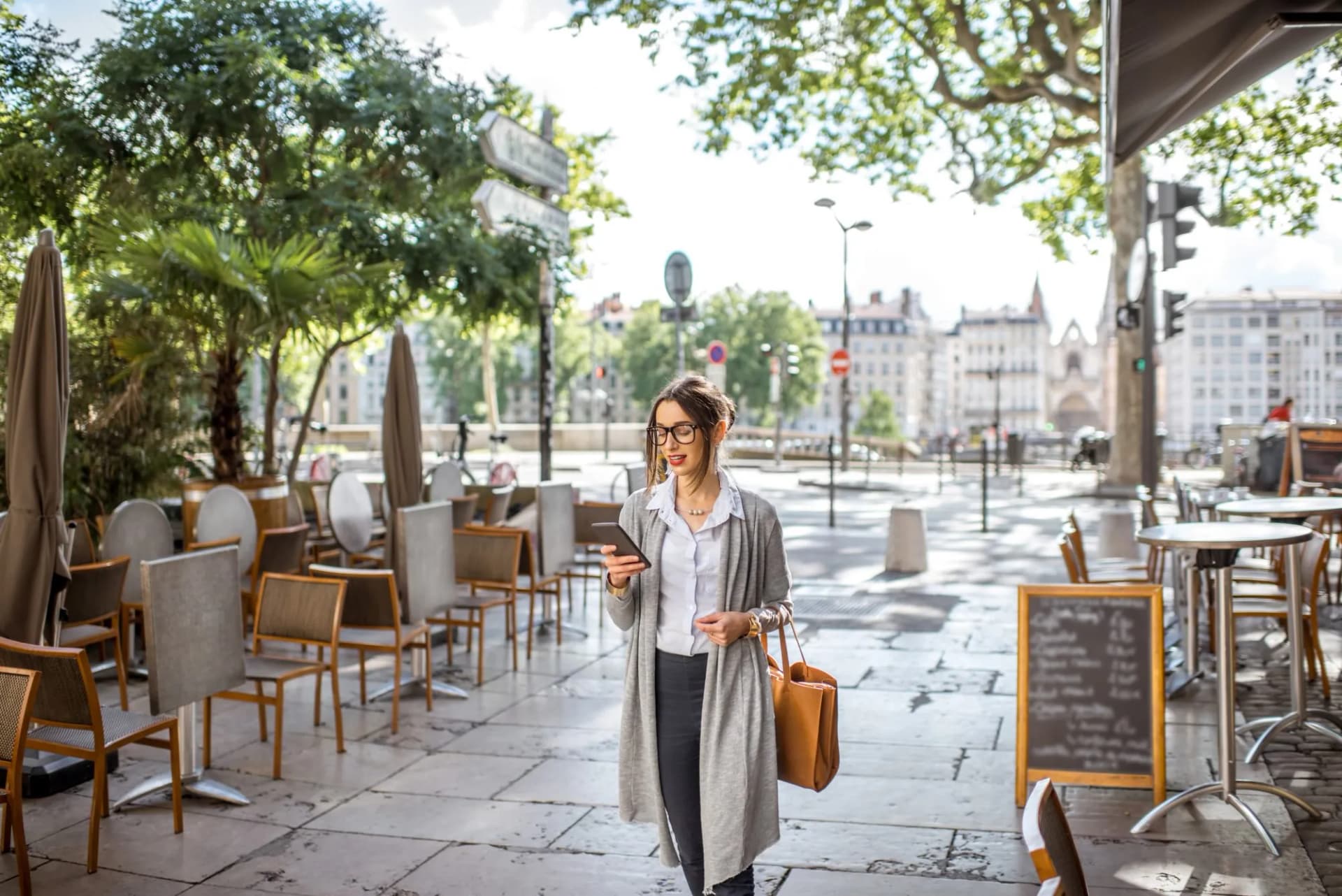 Stylish businesswoman walking with phone past empty outdoor cafe seating in Lyon Old Town.