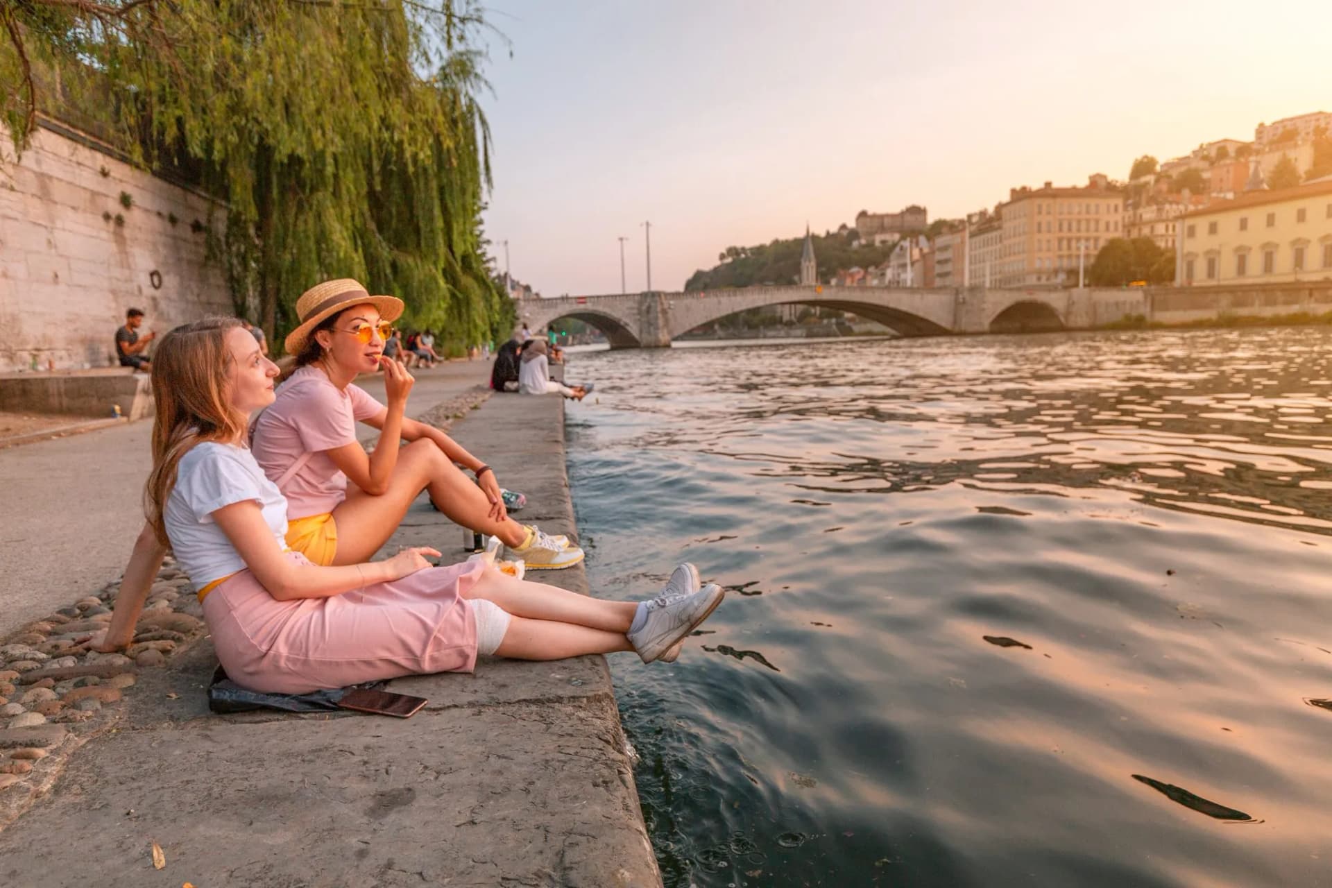 Two friends relaxing by the River Saône with a bridge and Lyon cityscape at sunset