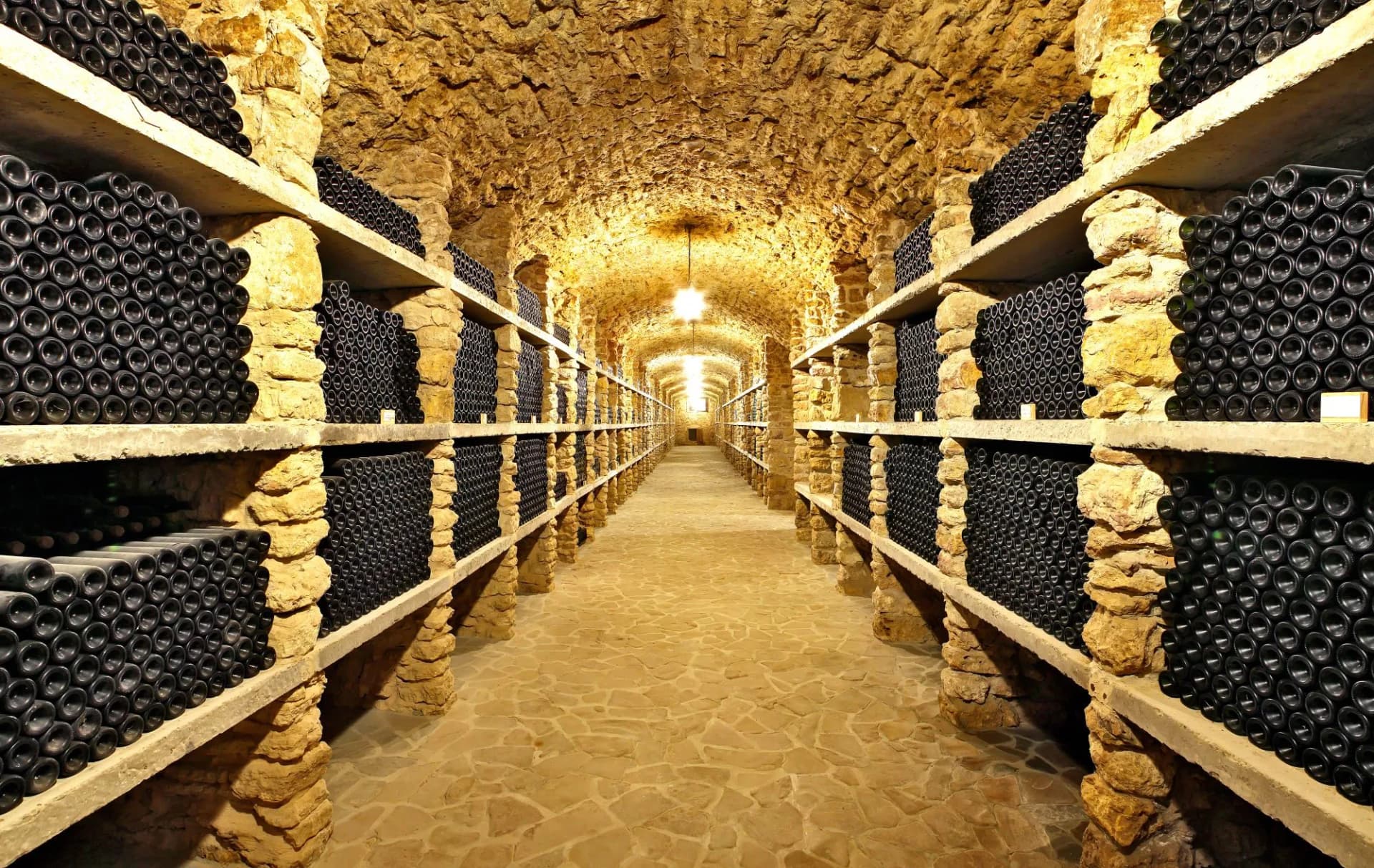 Wine bottles stacked on stone shelves in a long, illuminated ancient cellar tunnel