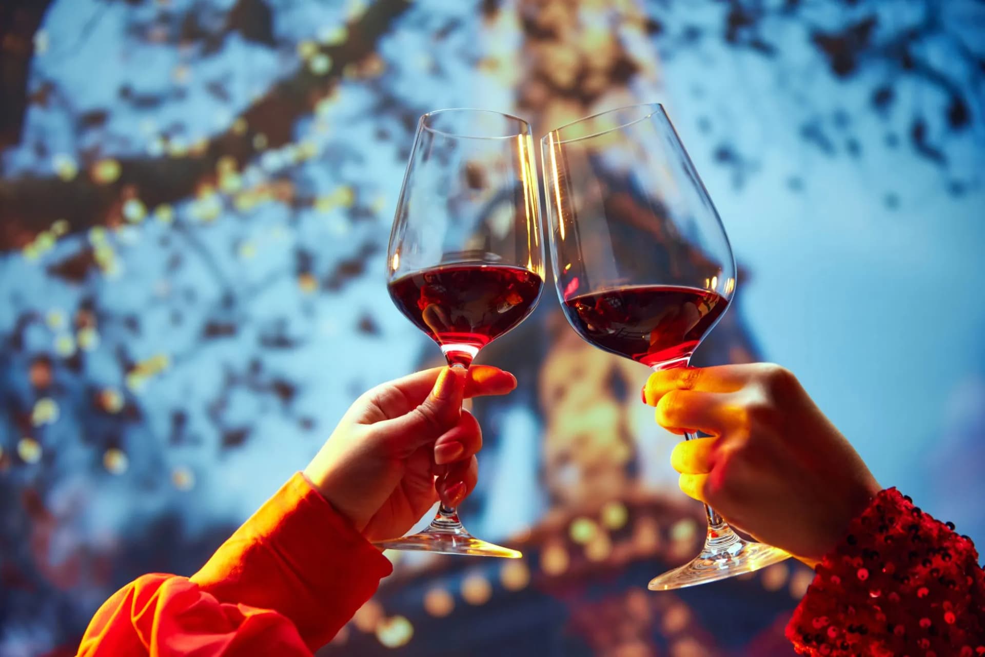 Two hands toast with red wine glasses, blurred Eiffel Tower and lights in background.