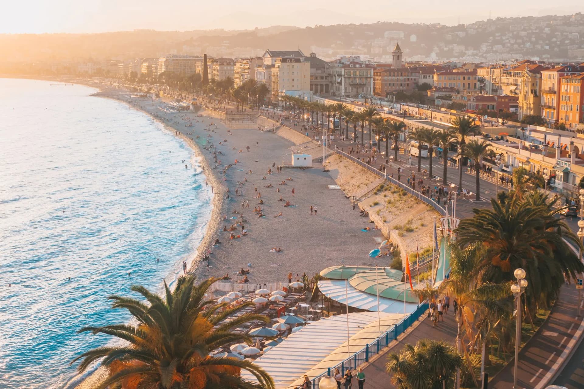 Beachgoers relaxing on pebble beach next to palm-lined promenade in Nice at sunset