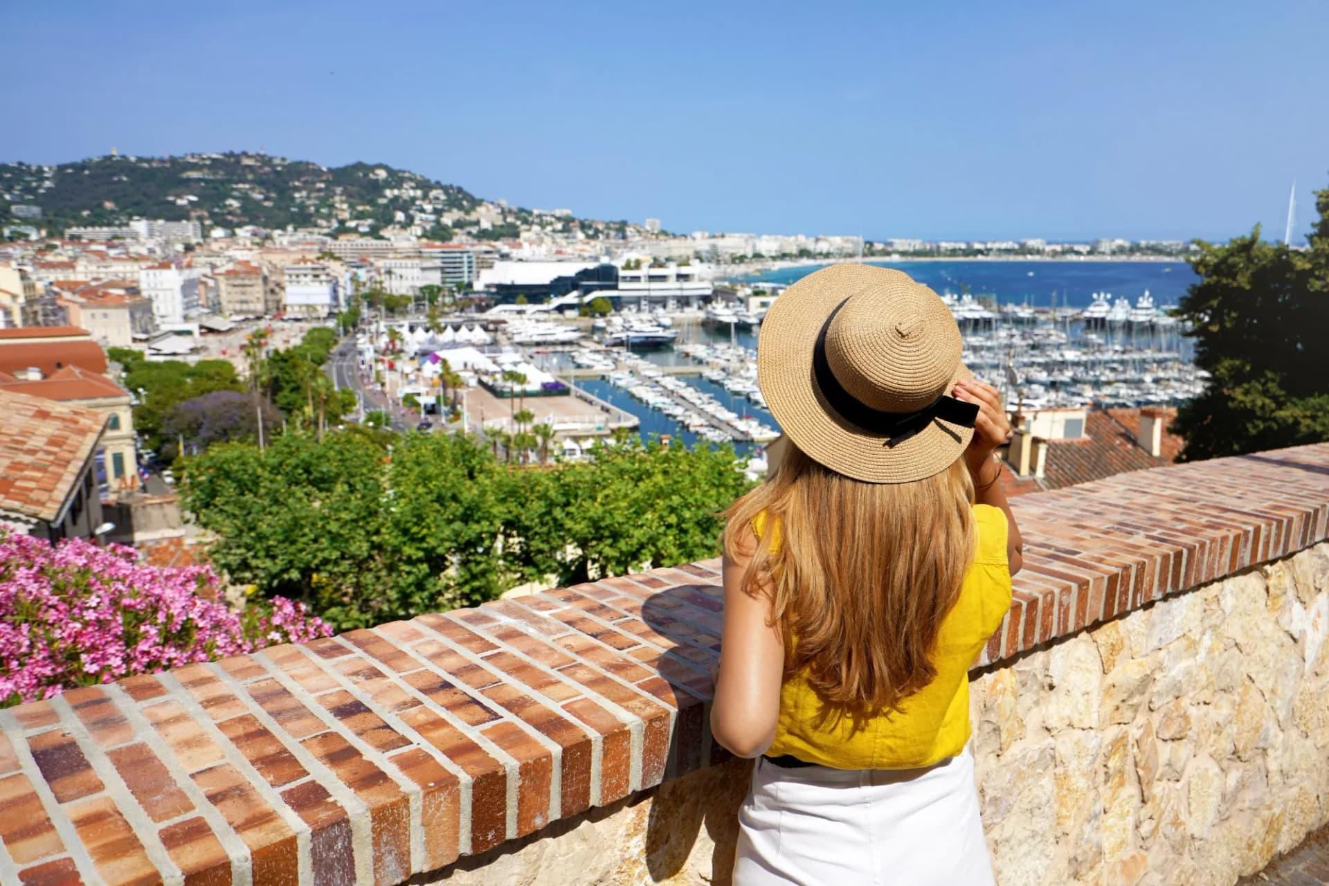 Woman in straw hat viewing cityscape and marina in Cannes, France from a stone wall.