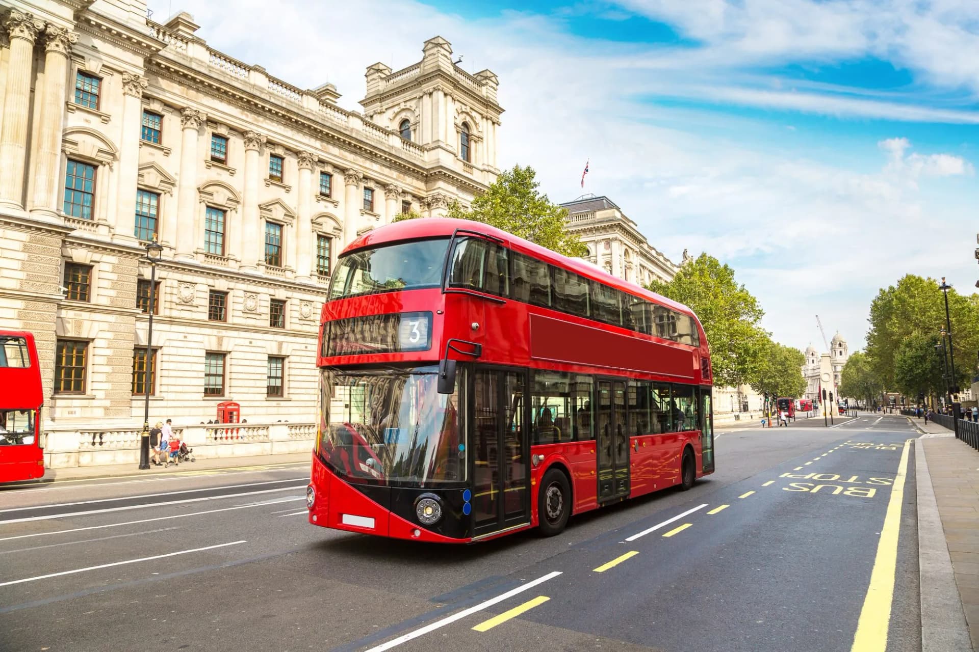 Modern red double-decker bus driving past classical building in London.