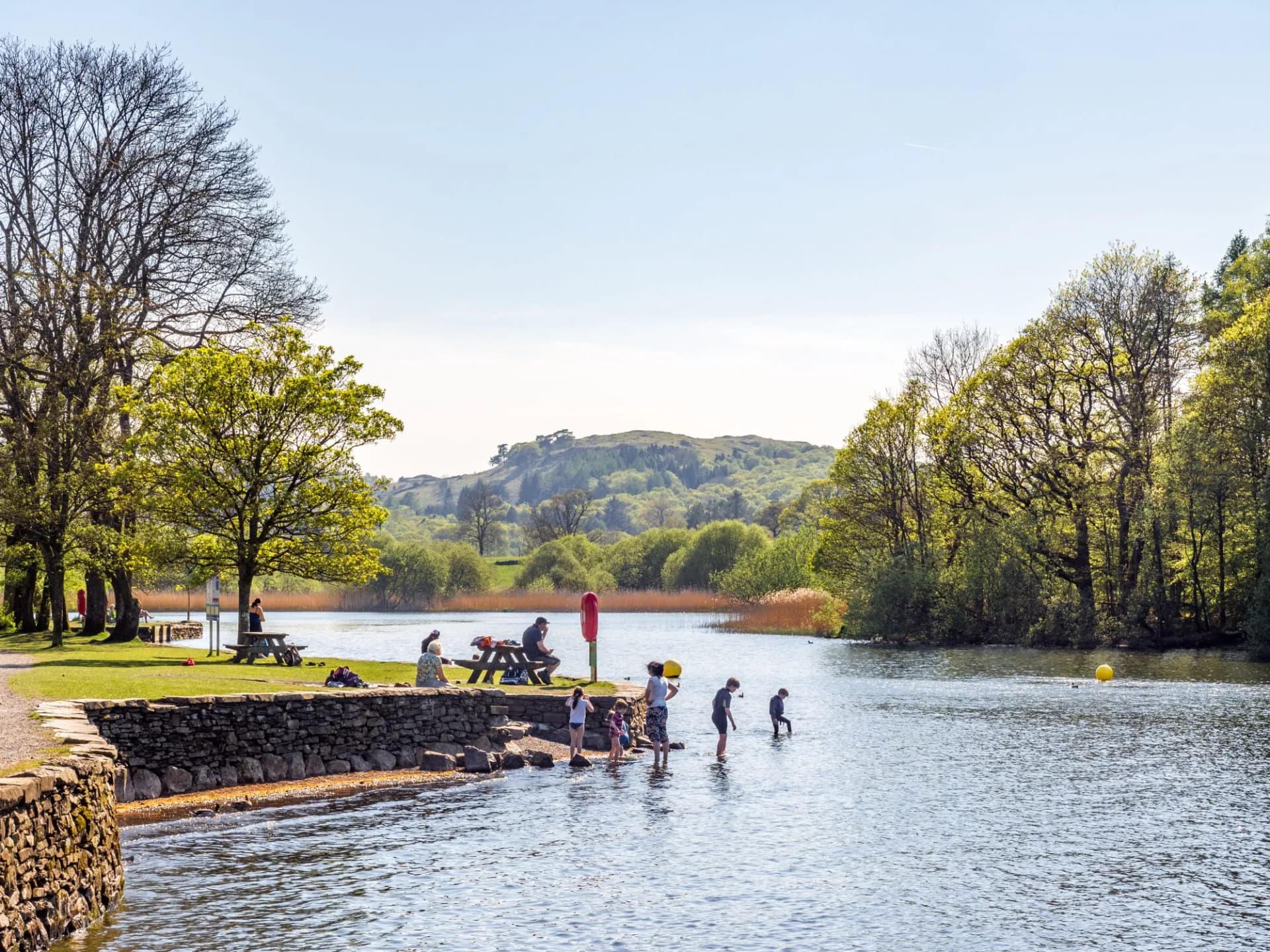 Tourists cooling off in Lake Windermere at Fell Foot on a sunny day with green hills.