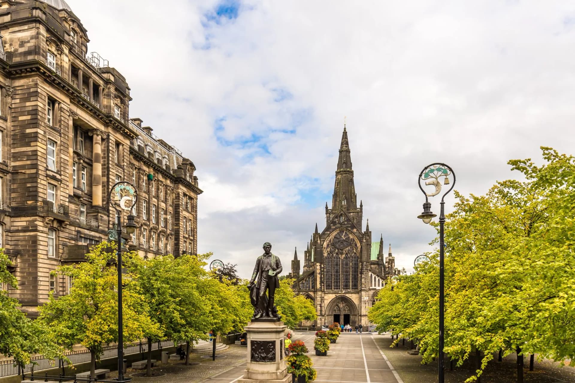 Statue and ornate church spire in George Square, Glasgow, Scotland, framed by trees.