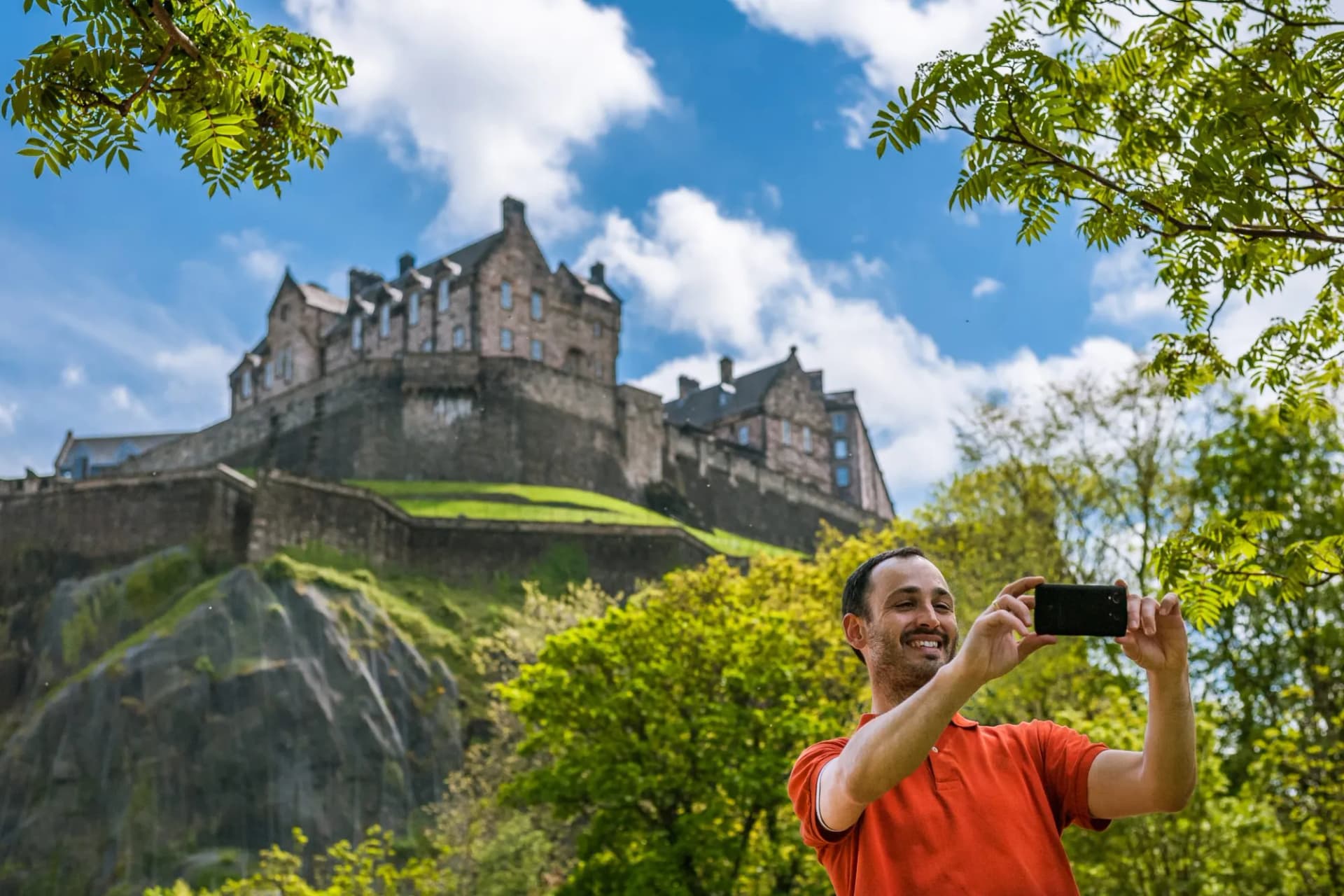 Tourist taking a selfie with Edinburgh Castle on a sunny day with green trees.