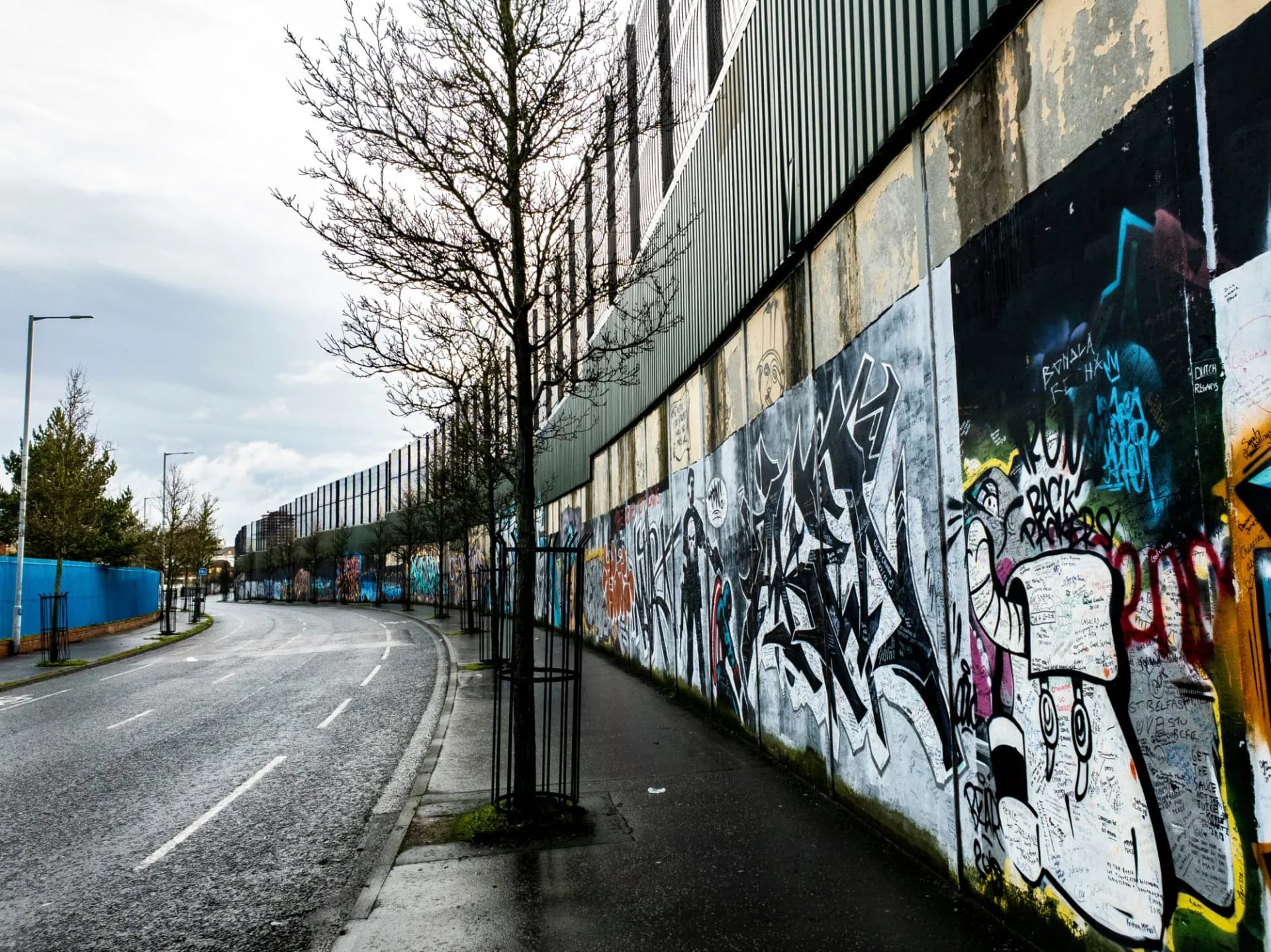 Peace Wall in Belfast with extensive graffiti and bare trees along a wet road.