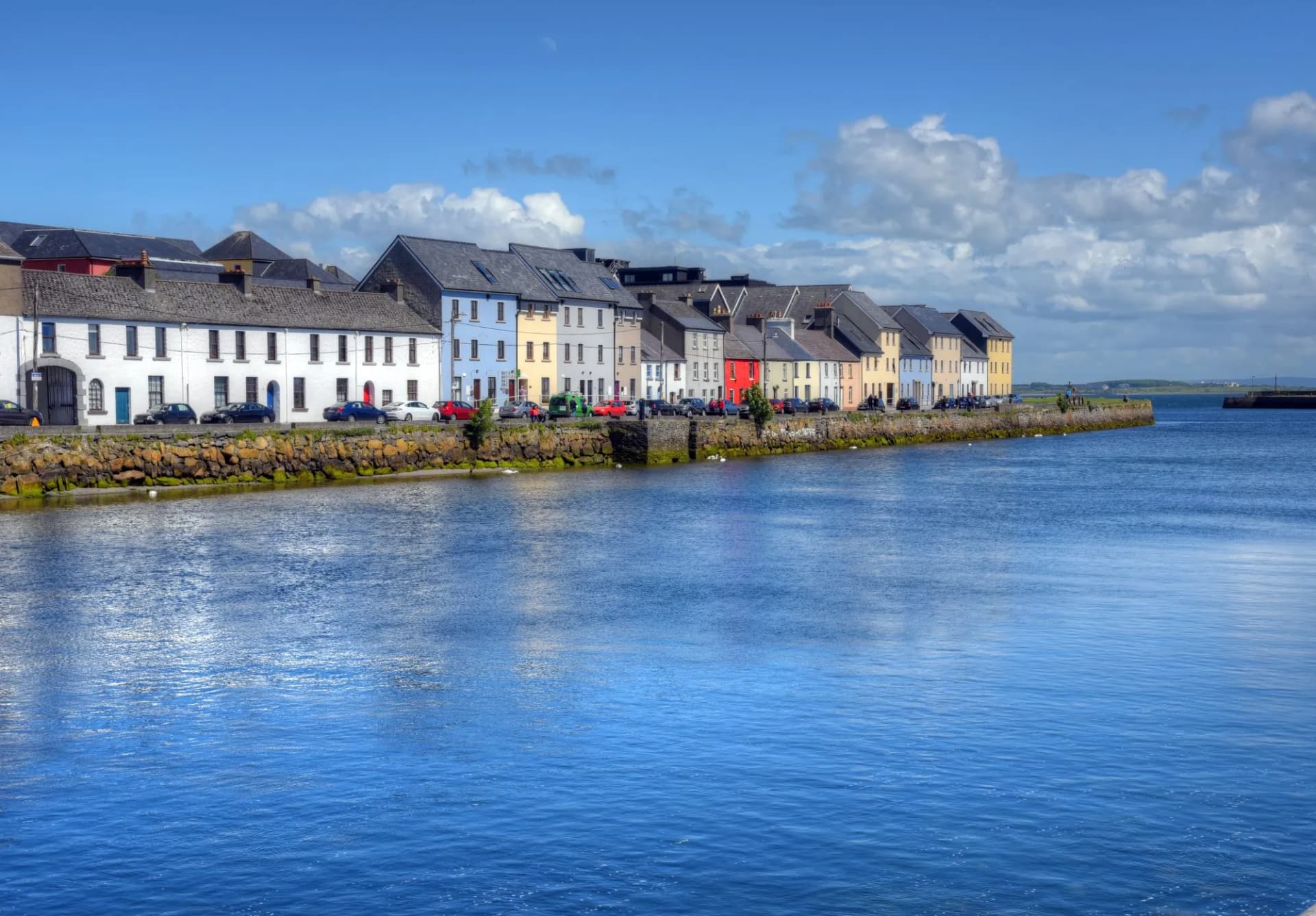 Colorful waterfront buildings along a stone quay in Galway, Ireland, under a blue sky.