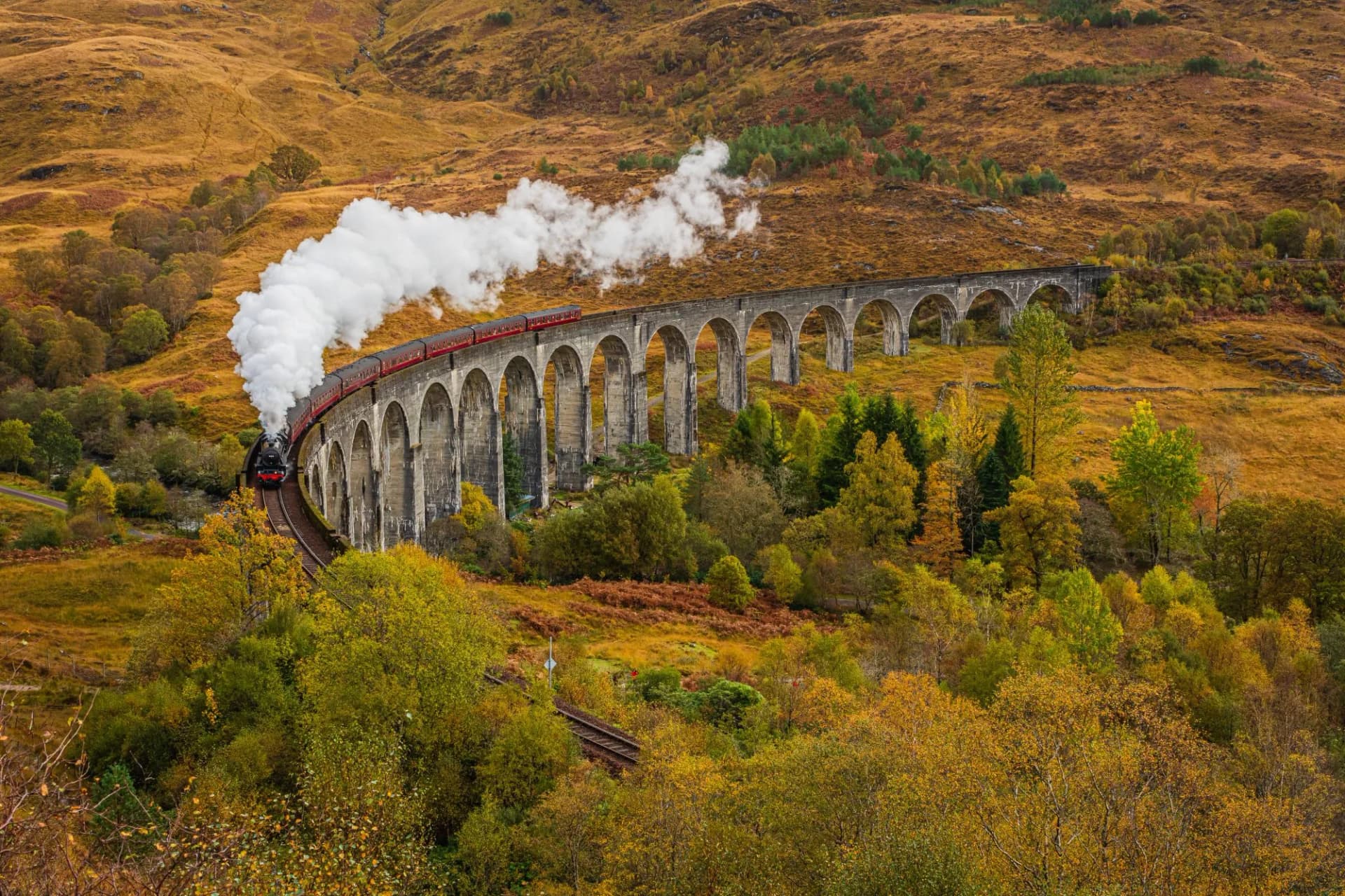 Steam train crossing Glenfinnan Viaduct in Scotland with autumn foliage on hillsides.