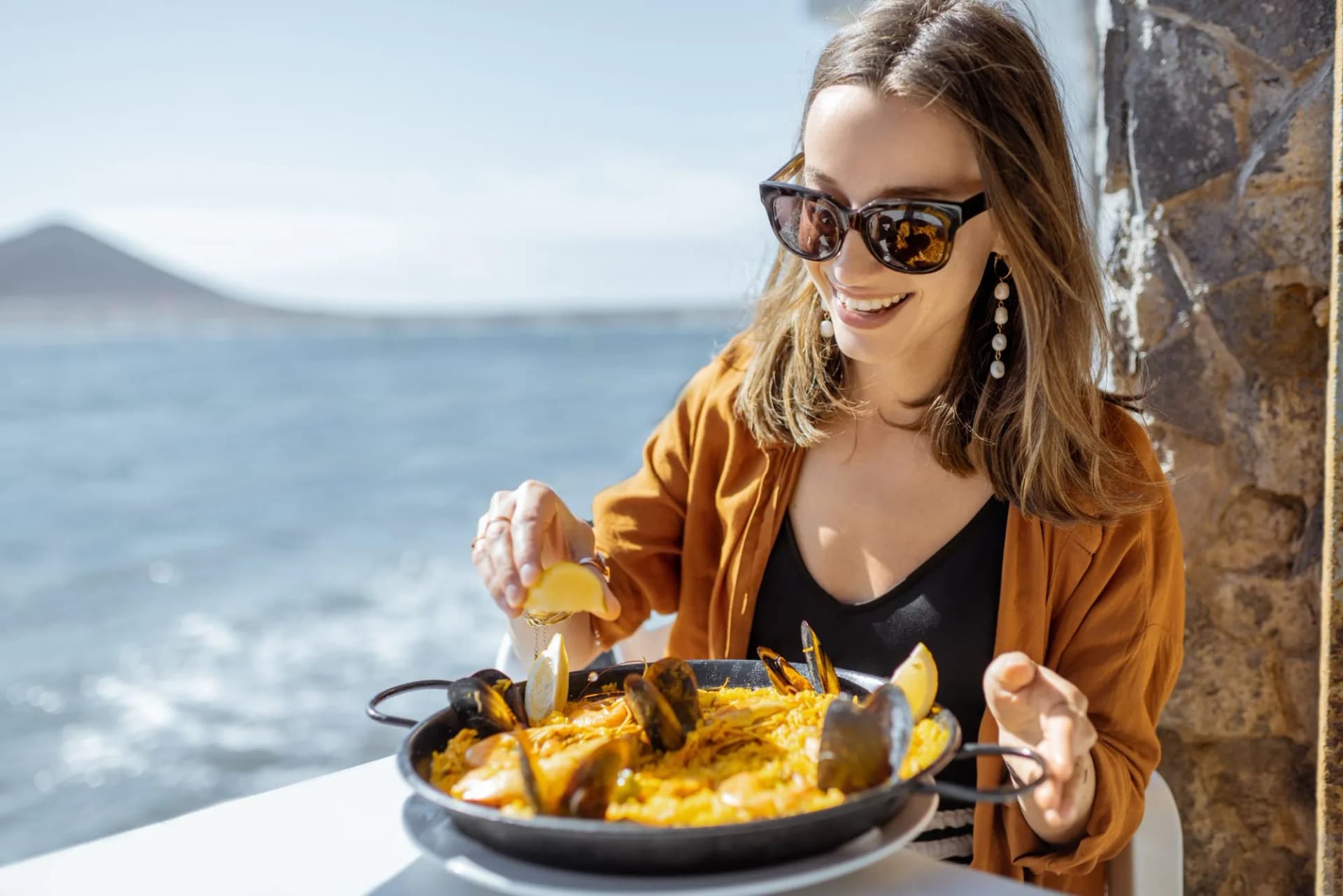 Woman eating seafood paella on restaurant terrace near ocean with distant mountain.