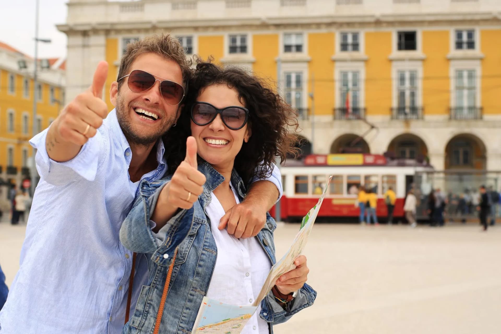 Young couple tourists giving thumbs up with map in Lisbon square with yellow building and tram.