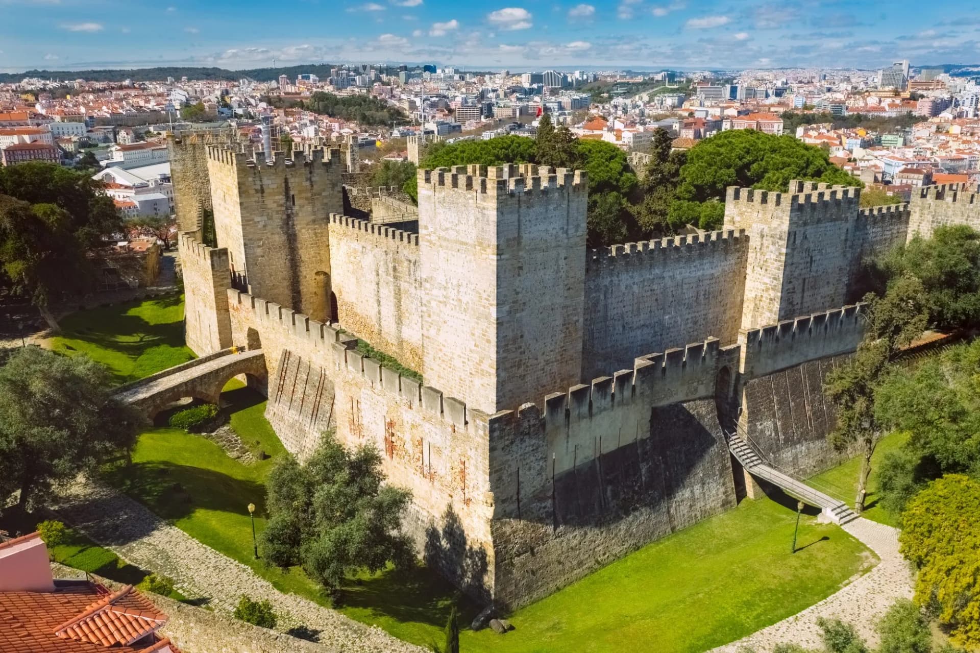 Aerial view of São Jorge Castle walls with green grounds and Lisbon city skyline in background.