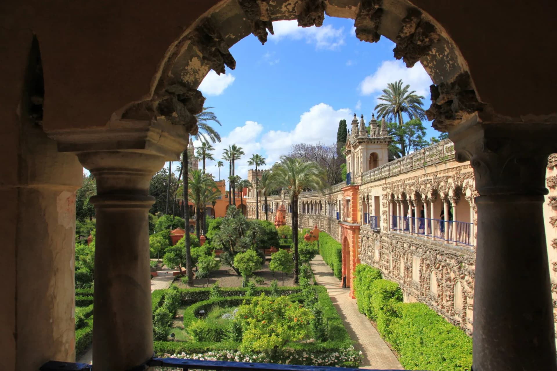View through an archway of lush gardens and ornate palace architecture in the Gardens of Alcazar, Seville, Spain.