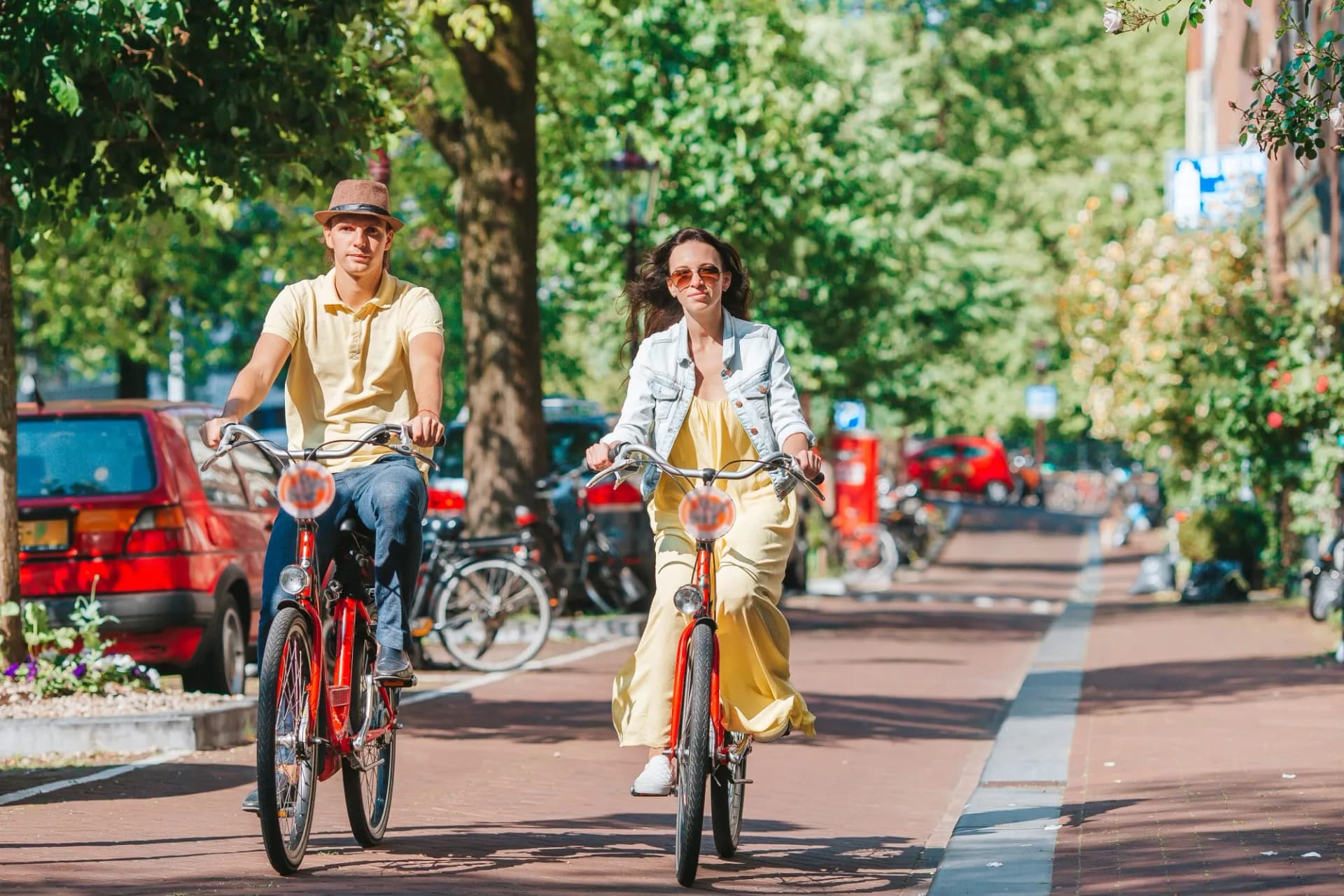 Couple cycling on bikes down a tree-lined street in Amsterdam on a sunny day.