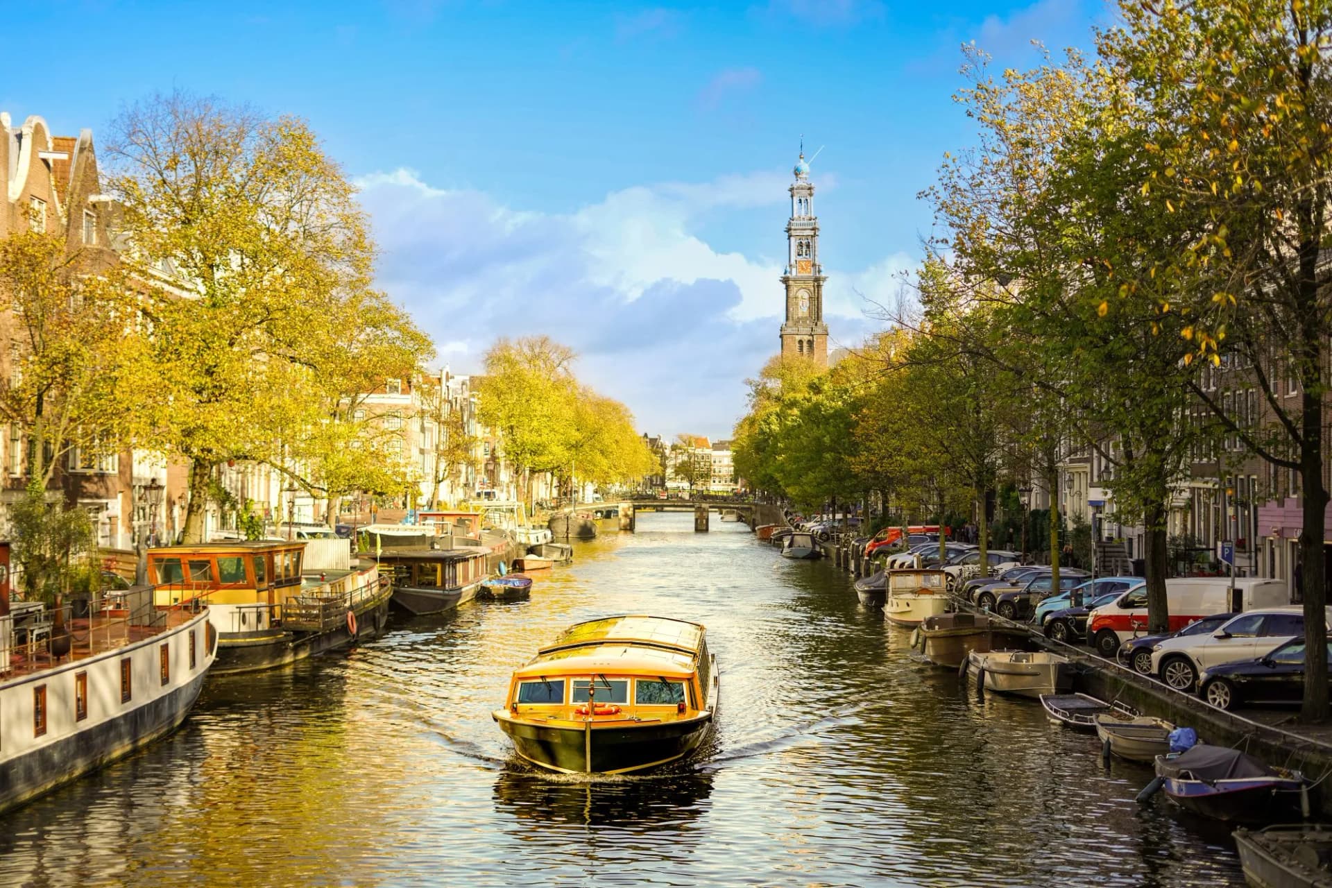 Cruise boat navigating Amsterdam canal with autumn trees and Westerkerk tower.