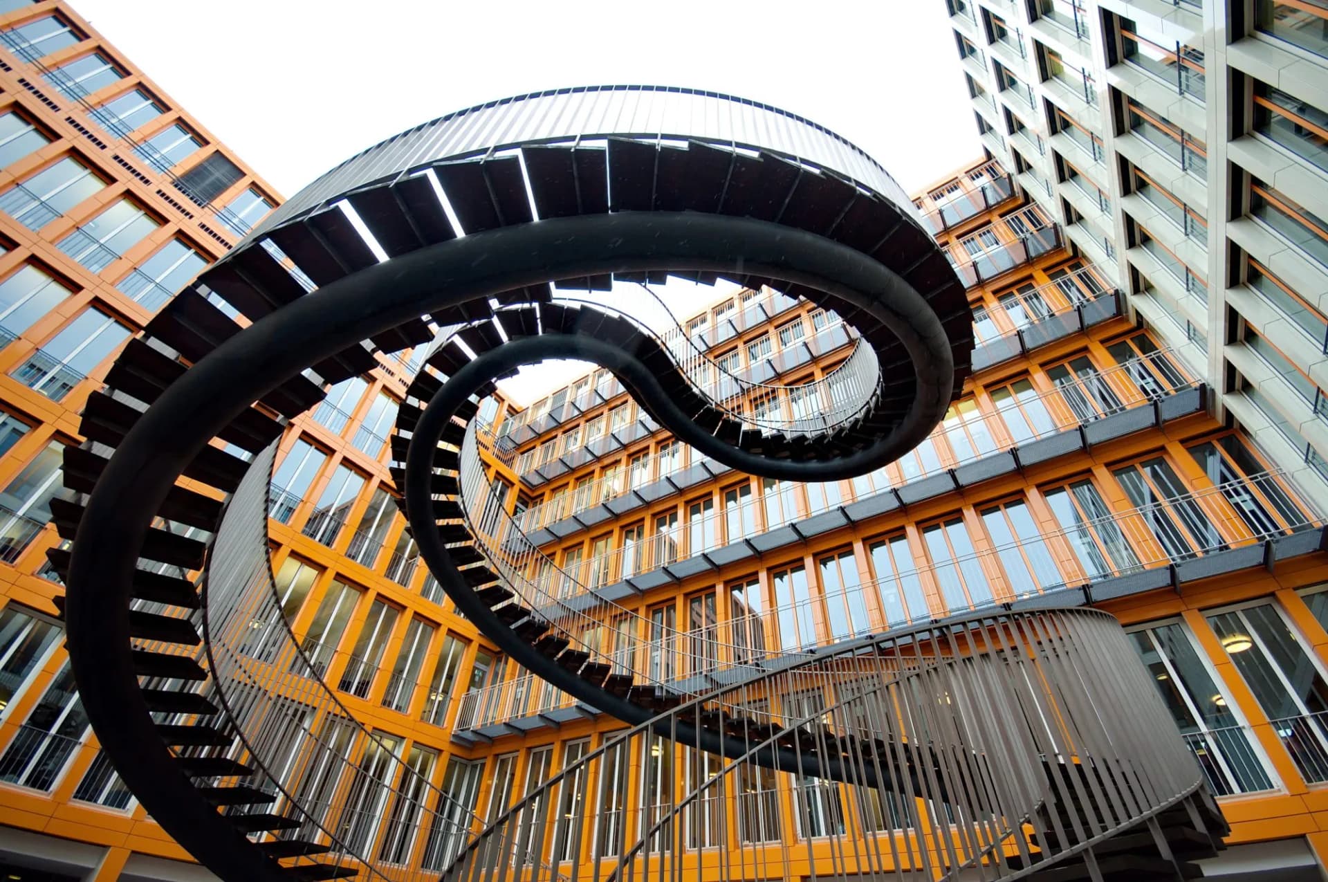Endless, swirling black steel stairway in courtyard of orange and white modern buildings in Munich, Germany.