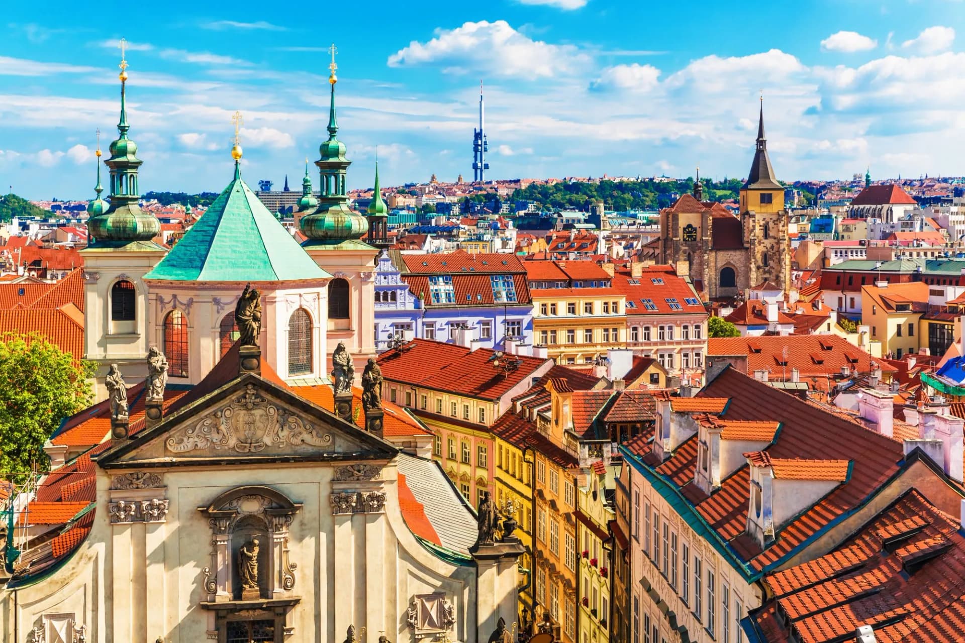 Aerial view of Prague, Czech Republic, showing historic buildings with red roofs and green spires under a blue sky.