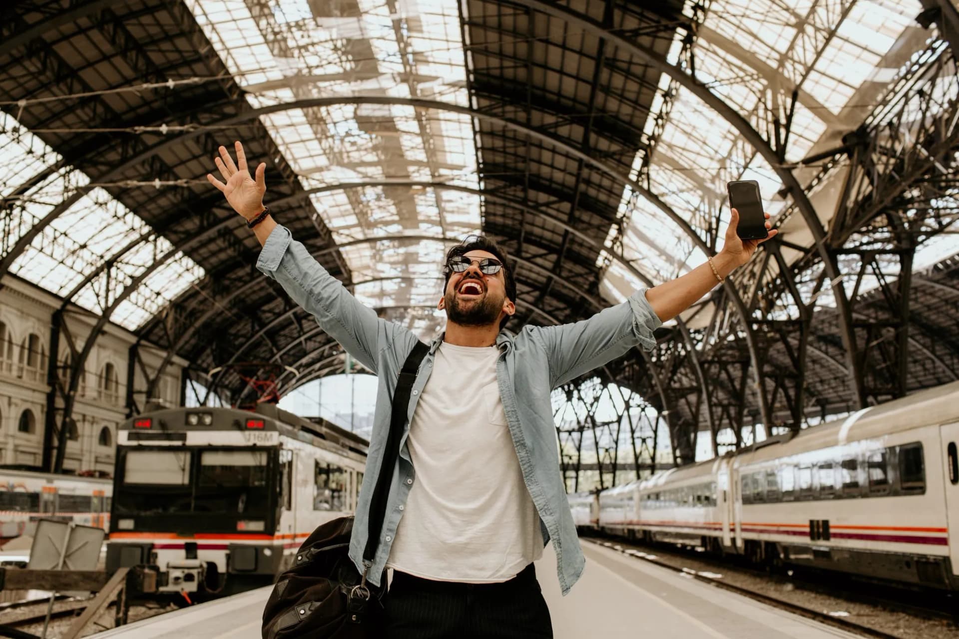 Happy traveler with arms raised holding smartphone at train station platform with trains.