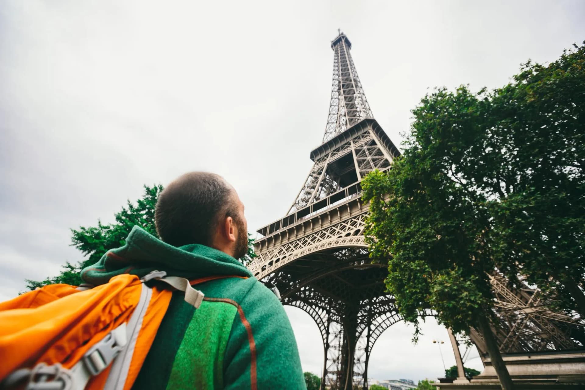 Tourist with backpack looking up at the Eiffel Tower in Paris under cloudy weather.