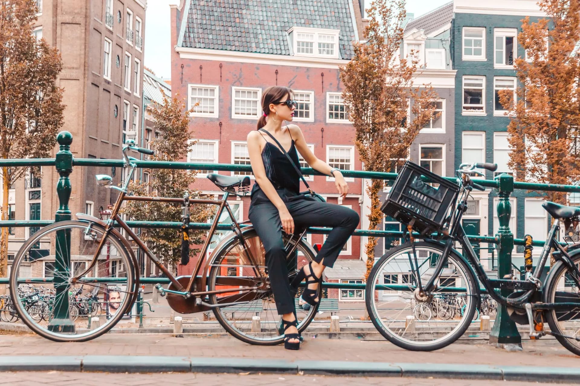 Woman sitting on bicycle on Amsterdam bridge with historic canal houses and autumn trees.