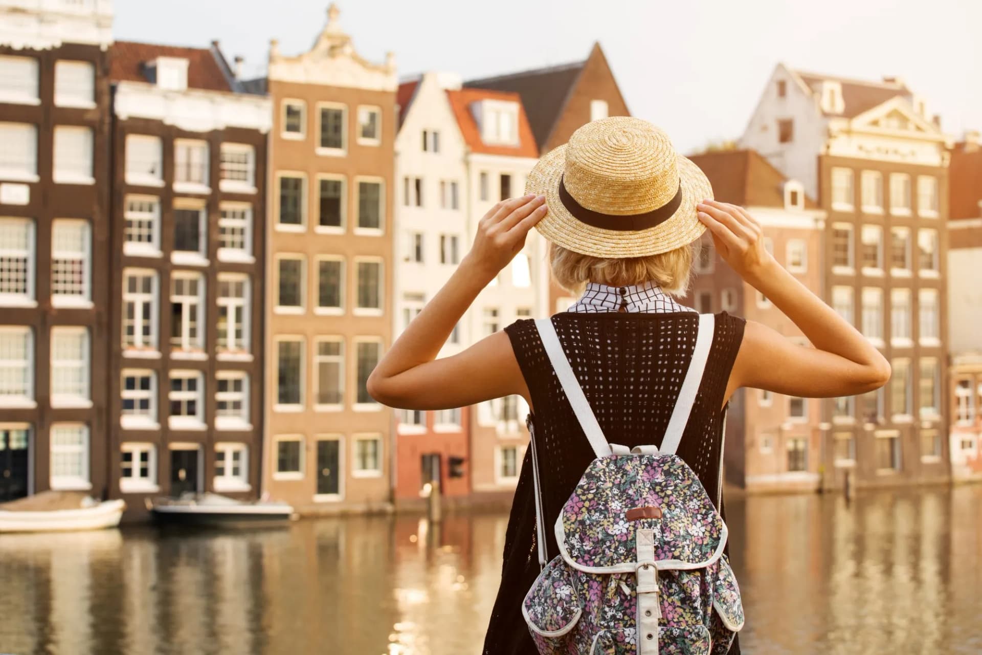 Woman with straw hat and floral backpack looking at canal houses in Amsterdam.
