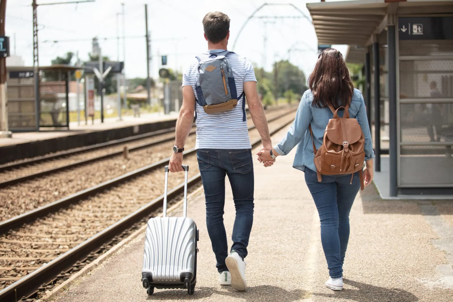 Couple walking with luggage on a train station platform beside the tracks.