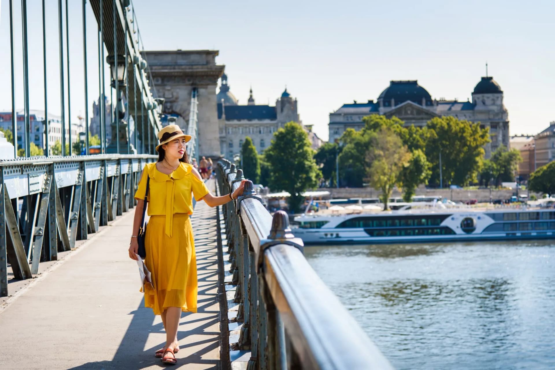 Woman walking on the Chain Bridge with Budapest buildings and river cruise ship visible.