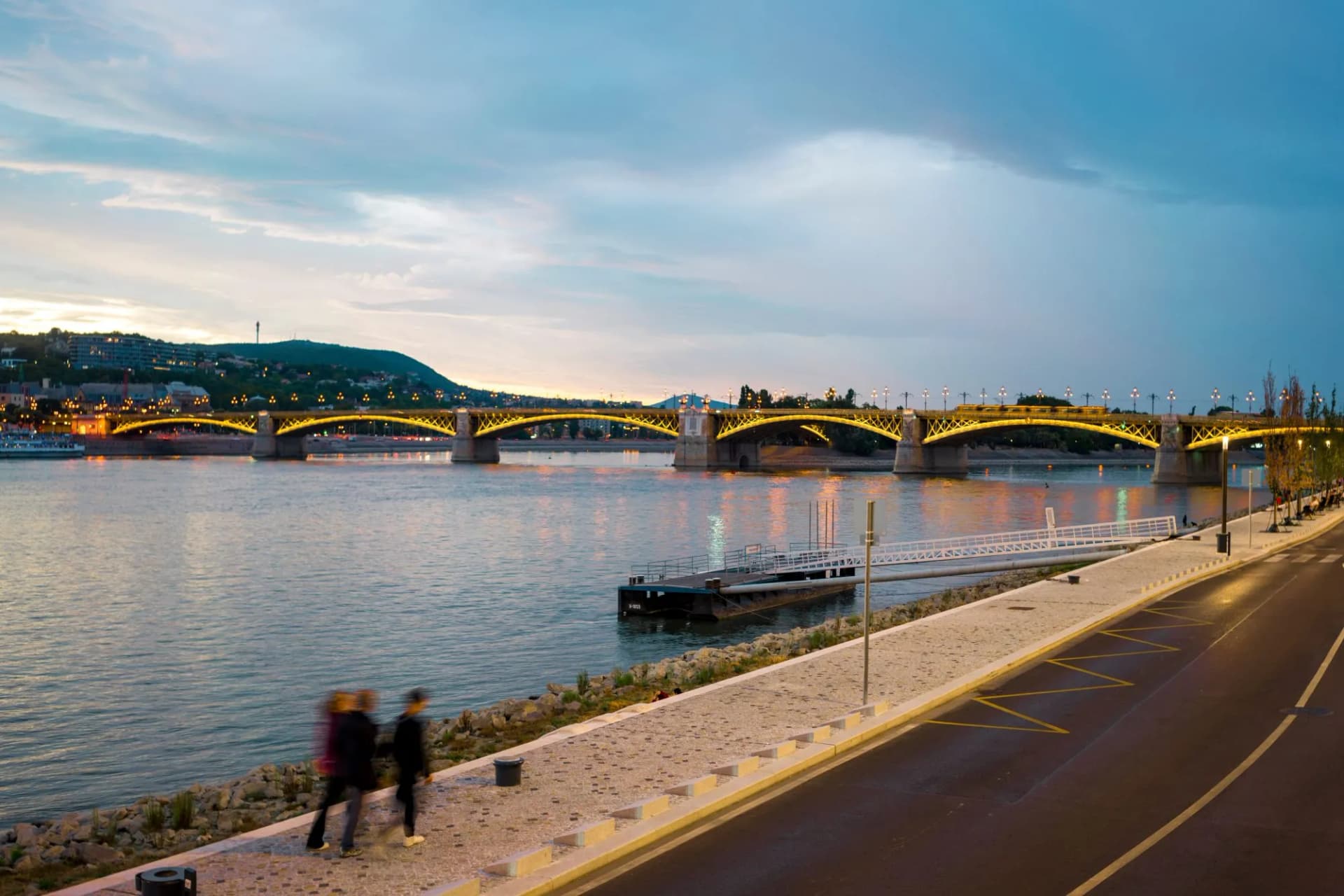 Illuminated Margit Bridge over the Danube River at dusk with people walking along the river bank in Budapest, Hungary.