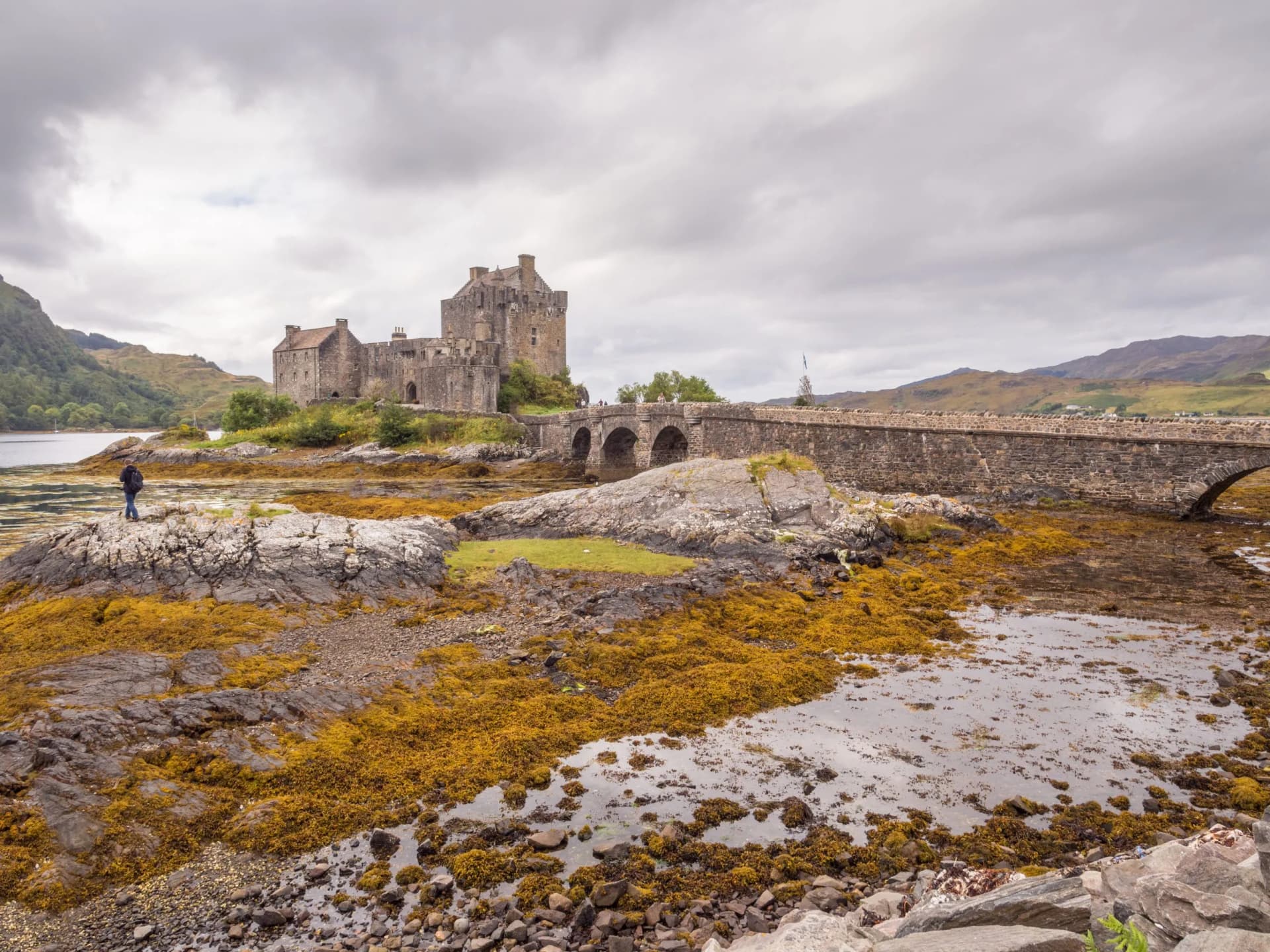 Eilean Donan Castle at low tide with stone bridge, mountains, and person on rocks in Kyle of Lochalsh, Scotland.