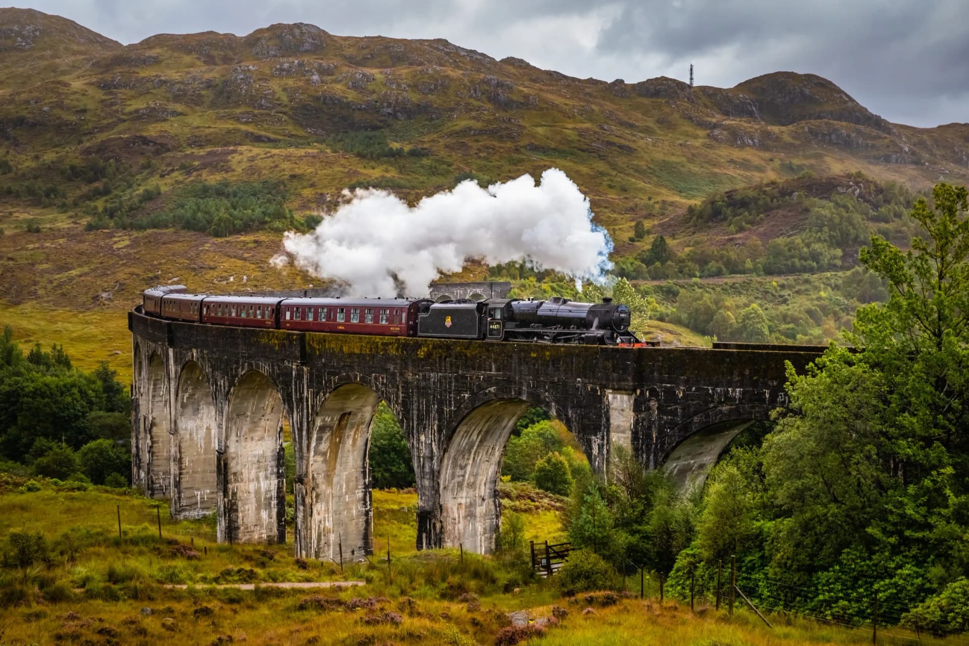 Steam train crossing Glenfinnan Viaduct with rolling green and brown hills under a cloudy sky.